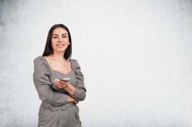 Resmi elbise giyen gülümseyen genç çekici iş kadını elinde akıllı telefon tutuyor. Arka planda boş beton duvar var. Hayal gücü kavramı ve yaratıcı fikirler için ilham.