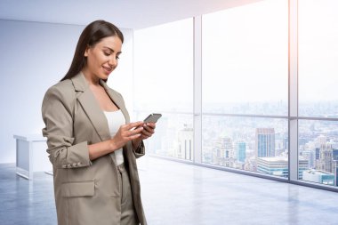 Ofisteki bej takım elbiseli kadın, panoramik pencereli iş odasının arka planında telefonla yazı yazıyor. İş ve teknoloji kavramı