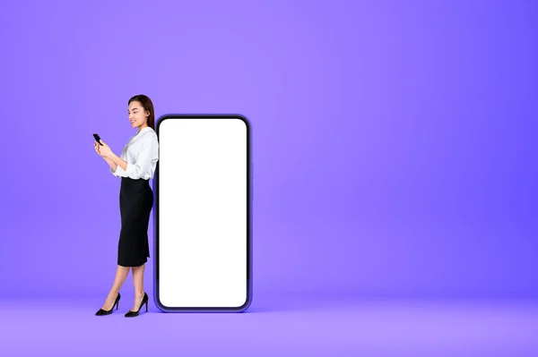 Smiling young attractive businesswoman wearing formal wear is typing on smartphone near empty mobile case offer mock up copy space. Purple background. Concept of imagination and inspiration