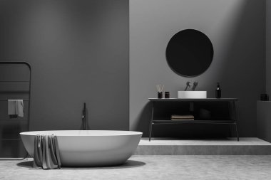 Beyaz küvetli, gri beton zeminli koyu renk banyo. Lavabo ve yuvarlak ayna, güvertede aksesuarlar, havlu demiryolu merdiveni. Mokup boş gri duvar, 3B görüntüleme