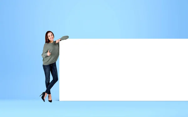 Woman gives like, thumb up, smiling near empty canvas. Mockup blank whiteboard on blue background. Concept of feedback and recommend