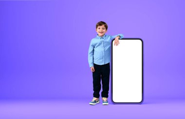 Çocuk gülümsüyor, uzun süre ayakta duruyor. Akıllı telefonda uzanıyor. Mor arka plan, boş ekran gibi. Sosyal medya ve çevrimiçi eğitim. Mobil uygulama kavramı