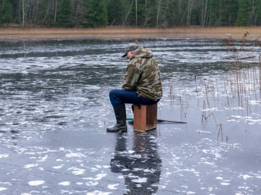 Buzda balık tutan balıkçı, buz yüzeyinde yansımalar, kıyıda çıplak ağaç siluetleri, ön planda buz dokusu.