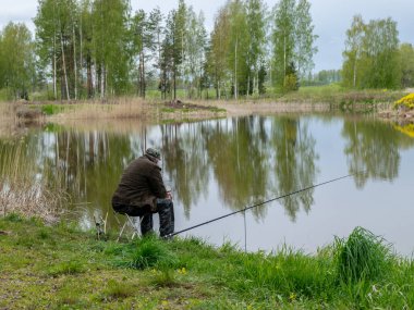 İlkbahar manzarası gölün kıyısında bir balıkçıyla, yeşil salonda balıkçı aksesuarları ön planda.