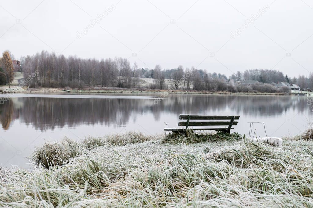 Foggy morning at dawn with forest by the lake, frosted wooden bench by the lake, late autumn