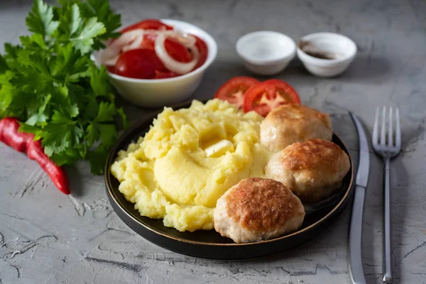 İçten bir öğle yemeği: patates püresi, domuz pirzolası, tabakta domates dilimleri, beyaz kasede domates salatası, gri arka planda taze yeşiller..
