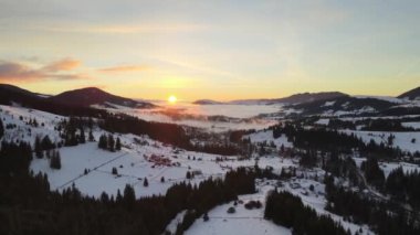 İnsansız hava aracı kışın dağ vadisinde uçuyor. Gün doğumunda çam ormanı tepelerinde çayırlarda kulübelerle kaplı kar manzarası.