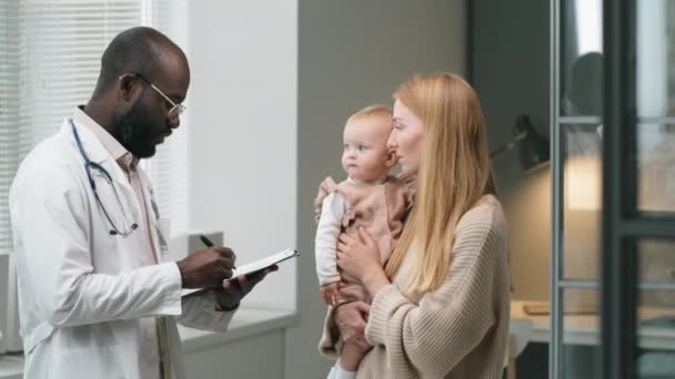 在诊所会诊期间 非裔美国医生在剪贴板上做笔记 并与抱着小女儿的白人妇女交谈 — 图库视频影像