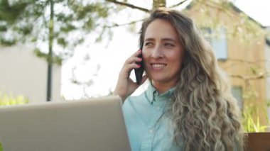 Düşük açılı güzel beyaz kadın yaz günü dizüstü bilgisayarıyla parkta oturuyor, gülümsüyor ve cep telefonuyla konuşuyor.