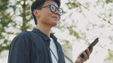 Asyalı genç bir kadının günlük kıyafetleri ve gözlükleriyle sokakta dikilip akıllı telefon kullanmasının düşük açılı görüntüsü.