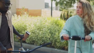Afrikalı genç bir adamın bisiklet ve kahveyle yürüyüp, beyaz bir kız arkadaşla parkta scooter sürerken görüntüsünü kaldır.