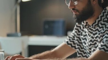 Yatay yan görüntü, sakallı, gözlüklü, dizüstü bilgisayarda çalışan genç bir adamın yavaş çekim çekimi.