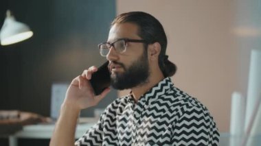 Gözlüklü yakışıklı Orta Doğulu bir adamın ofisinde oturmuş telefonla konuşurken çekilmiş yatay görüntüsü.
