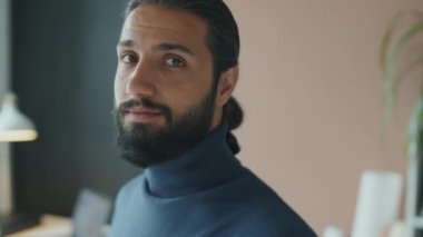 Yüzü sakallı, uzun saçlı, balıkçı yaka kazaklı ve kameraya bakan genç Orta Doğulu bir adamın yatay yakın plan portresi.