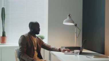Genç, Afro-Amerikan bir adamın ofiste oturup dizüstü bilgisayardan bir şey izlerken çekilmiş yatay portresi.