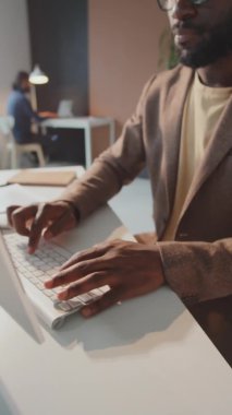 Dikey yavaş çekim, yakışıklı siyahi adamın bilgisayar ekranının önünde oturup işini yaparken ki görüntüsü.