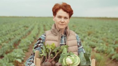 Beyaz kadın portresi elinde taze lahana ve pancar dolu kasayla tarlada kameraya poz veriyor.