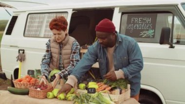 Afro-Amerikalı erkek ve beyaz kadın, yerel çiftlik ürünleri satarken minibüsün önüne taze sebze koyuyorlar.