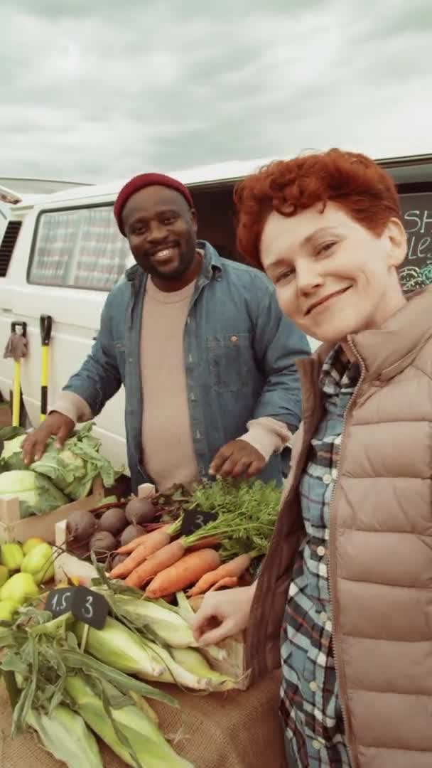 若い白人女性とアフリカ系アメリカ人男性の垂直ショットは 屋外市場で農場から新鮮な野菜を販売しながら オンラインビデオ通話を介してカメラに手を振って話しています — ストック動画
