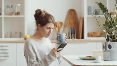 Kafkasyalı genç kadın fincandan çay içiyor, akıllı telefondan mesaj atıyor ve yemek masasında otururken gülümsüyor ve çiçekleri suluyor.