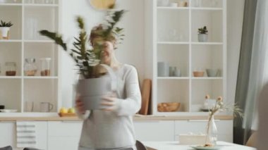 Kablosuz kulaklık takan, elinde gülen yüzle dekore edilmiş ev bitkisi tutan, ona bakıp dans eden ve kendini izolasyonda eğlendiren genç, pozitif bir kadın.