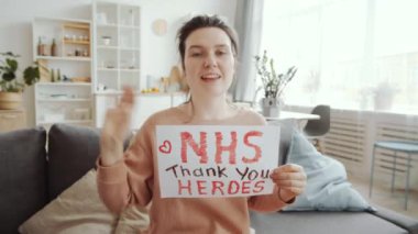 Elinde NHS yazılı kağıt tutan genç, pozitif bir kadın siz kahramanlara teşekkür ediyor. Evdeki kanepede otururken kameraya el sallıyor ve bir şeyler anlatıyor. Coronavirüs salgını kavramı