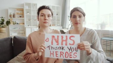 Elinde NHS yazılı kağıt olan iki bayan arkadaş kameraya bakıp evdeki kanepede otururken doktorlara teşekkür eden kahramanlara teşekkür ediyor. Covid-19 salgın konsepti