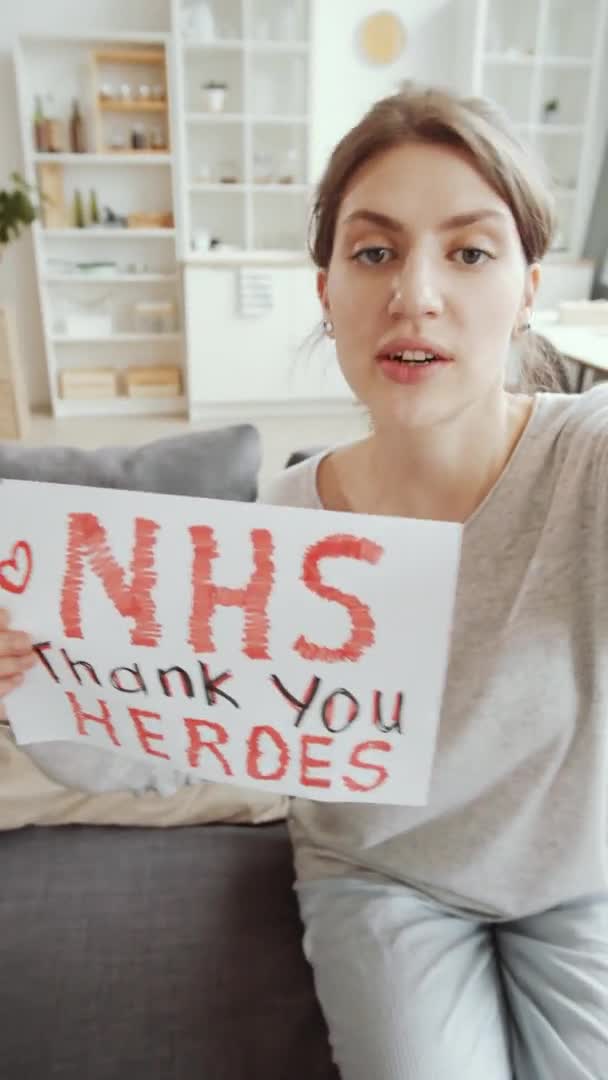 年轻漂亮女人拿着印有Nhs字样的纸片的垂直照片 感谢英雄的题词 看着相机 并在家里沙发上向医院的工作人员致谢 — 图库视频影像