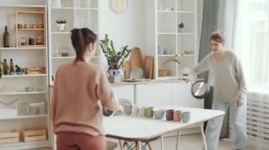 Evde izole bir şekilde vakit geçirirken mutfakta ağ yerine tava ve çay fincanı gibi tavalarla masa tenisi oynayan iki genç kadın.