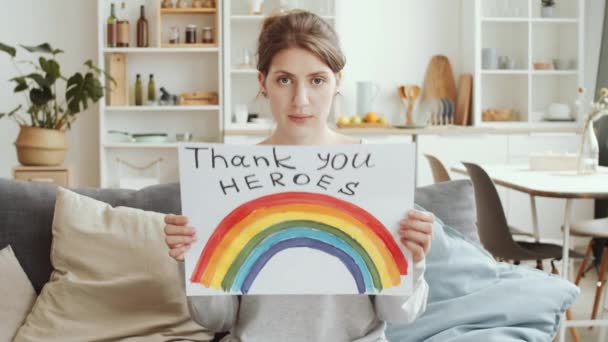 年轻的白人妇女展示手工制作的海报彩虹和Nhs感谢英雄的题词 并在验尸期间坐在沙发上看着相机 — 图库视频影像