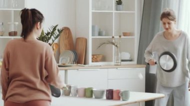 Mutfak masasında pinpon oynarken evde karantina altında kalmak yerine kızartma tavalarını raket ve çay kupası olarak kullanan iki genç ve neşeli kadın.