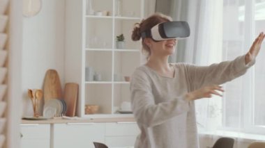 VR kulaklıklı genç, neşeli bir kadın evde tek başına otururken video oyunu oynarken gülümsüyor ve havaya dokunuyor.
