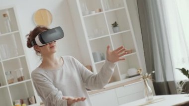 VR kulaklıklı genç beyaz kadın, karantina sırasında evinde video oyunu oynarken elinde görünmez bir nesne, havaya dokunuyor.