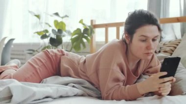 Kafkasyalı genç bir kadının rahat kıyafetleri içinde yatağında yatarken ve akıllı telefonuyla internette dolaşırken çekilmiş bir fotoğrafı.