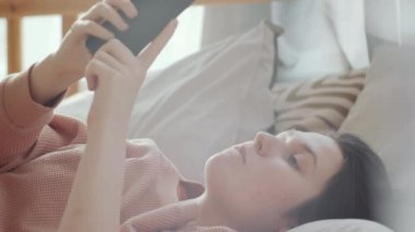 Genç esmer kadının yatakta yattığını ve tecrit sırasında evde otururken akıllı telefonuyla internete baktığını yakın plan çek.