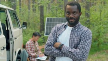 İşaretli tişört giyen genç Afro-Amerikan adamın yatay portresi diz üstü bilgisayarda çalışırken kollarını kavuşturup elektrik için güneş pili paneli kullanırken çekilmiş.