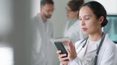 Laboratuvar önlüklü güzel bir bayanın, arka planda iki meslektaş konuşurken akıllı telefon ekranında görüntüsü.