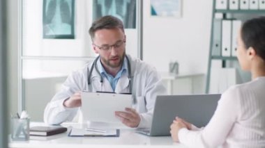 Laboratuar önlüklü, boynunda steteskop olan bir erkek doktor klinikte masasında oturuyor, kadın hastadan tıbbi geçmişini alıyor ve ona danışmanlık yapıyor.