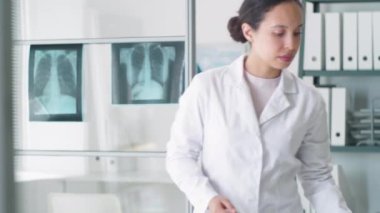 Laboratuvar önlüklü güzel, melez bir kadın doktor elinde not panosu, tıp ofisine gidiyor, masada oturuyor ve dizüstü bilgisayarla çalışıyor.