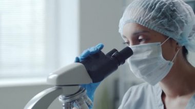 Tek kullanımlık şapkalı, steril eldivenli ve tıbbi maskesiyle laboratuarda çalışırken mikroskoptan bakan kadın bilim adamının göğüs görüntüsü.