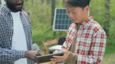 Genç Afro-Amerikan ve Asyalı bilim insanlarının dijital tablet ekranına bakarak ve sorunları tartışarak ormanda birlikte çalışmalarının yatay görüntüsü.