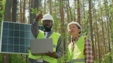Sarı yelekli ve beyaz şapkalı Afrikalı Amerikalı ve Asyalı iş arkadaşlarının güneş panelinin karşısında durup dizüstü bilgisayarlarını tutarak bir şeyler tartıştıkları düşük açılı hareket.
