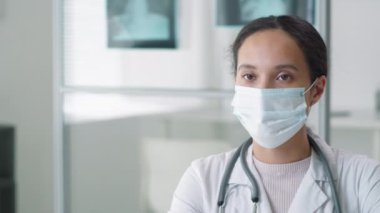 Tıbbi maske takmış esmer kadın doktor, laboratuvar önlüğü ve steteskop boynundayken klinikte otururken kameraya bakarken göğsünü çek.