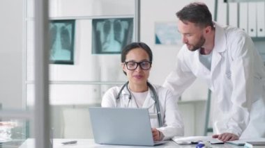 Laboratuvar önlüklü profesyonel Kafkas erkek doktor ve onun güzel melez bayan meslektaşı kameraya bakıp gülümserken aynı anda tıp ofisinde dizüstü bilgisayarda birlikte çalışıyorlar.