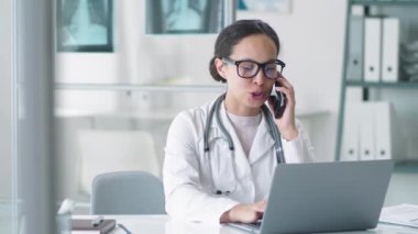 Laboratuar önlüklü, gözlüklü ve steteskoplu genç melez kadın doktor. Laptop kullanıyor, gülümsüyor ve klinikteki çalışma gününde cep telefonuyla konuşuyor.