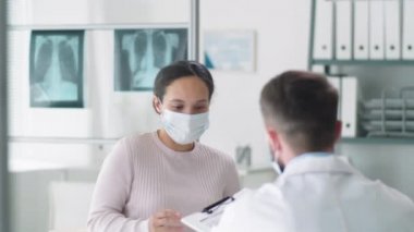 Koruyucu maske takan genç esmer kadın sağlık odasına giriyor, masada oturuyor, erkek doktora dosya veriyor ve onunla konsültasyonda konuşuyor. Covid-19 salgın konsepti