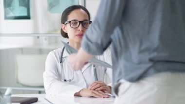 Laboratuvar önlüklü melez bir kadın doktor. Masasında oturmuş hamlelerin röntgenine bakıyor ve klinikte tıbbi danışmanlık yaparken erkek hastayla konuşuyor.