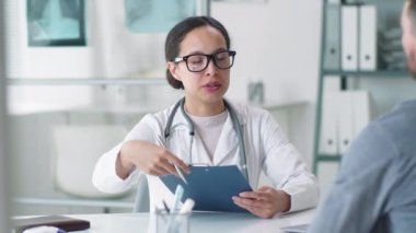 Laboratuvar önlüğü ve gözlüklü melez kadın doktor erkek hastayla konuşuyor ve klinikte muayeneden sonra imza vermesi için ona tıbbi hizmet anlaşması veriyor.