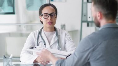 Laboratuvar önlüğü ve gözlüklü güzel kadın doktor konuşuyor. Erkek hastadan imzalı sağlık belgesi alıyor. Gülümsüyor ve muayenehanede çalışırken elini sıkıyor.