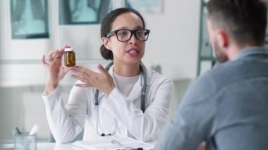 Laboratuvar önlüklü ve gözlüklü güzel bir kadın doktor erkek hastaya nasıl ilaç içileceğini ve ofisteki sağlık muayenesi sırasında ona haplarla dolu bir kap vereceğini söylüyor.
